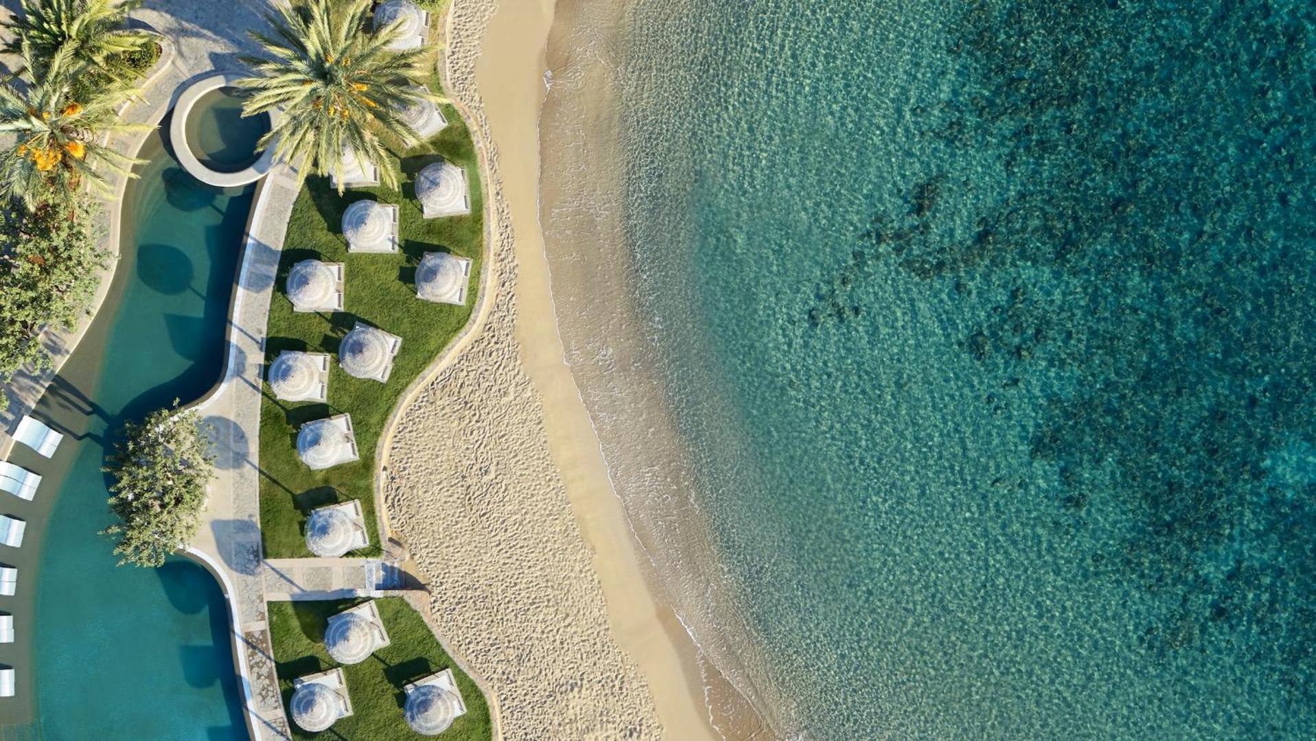Porto Elounda Golf & Spa Resort privātā pludmale no putna lidojuma — saulessargi uz zālāja un smiltīm pie kristāldzidra ūdens