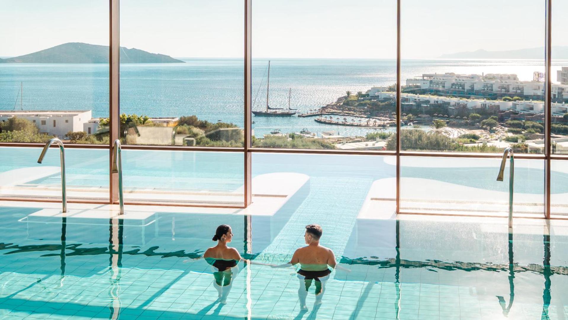 Porto Elounda Golf & Spa Resort iekštelpu baseins ar panorāmas logiem uz Mirabello līci Krētā un jahtu piekrastē