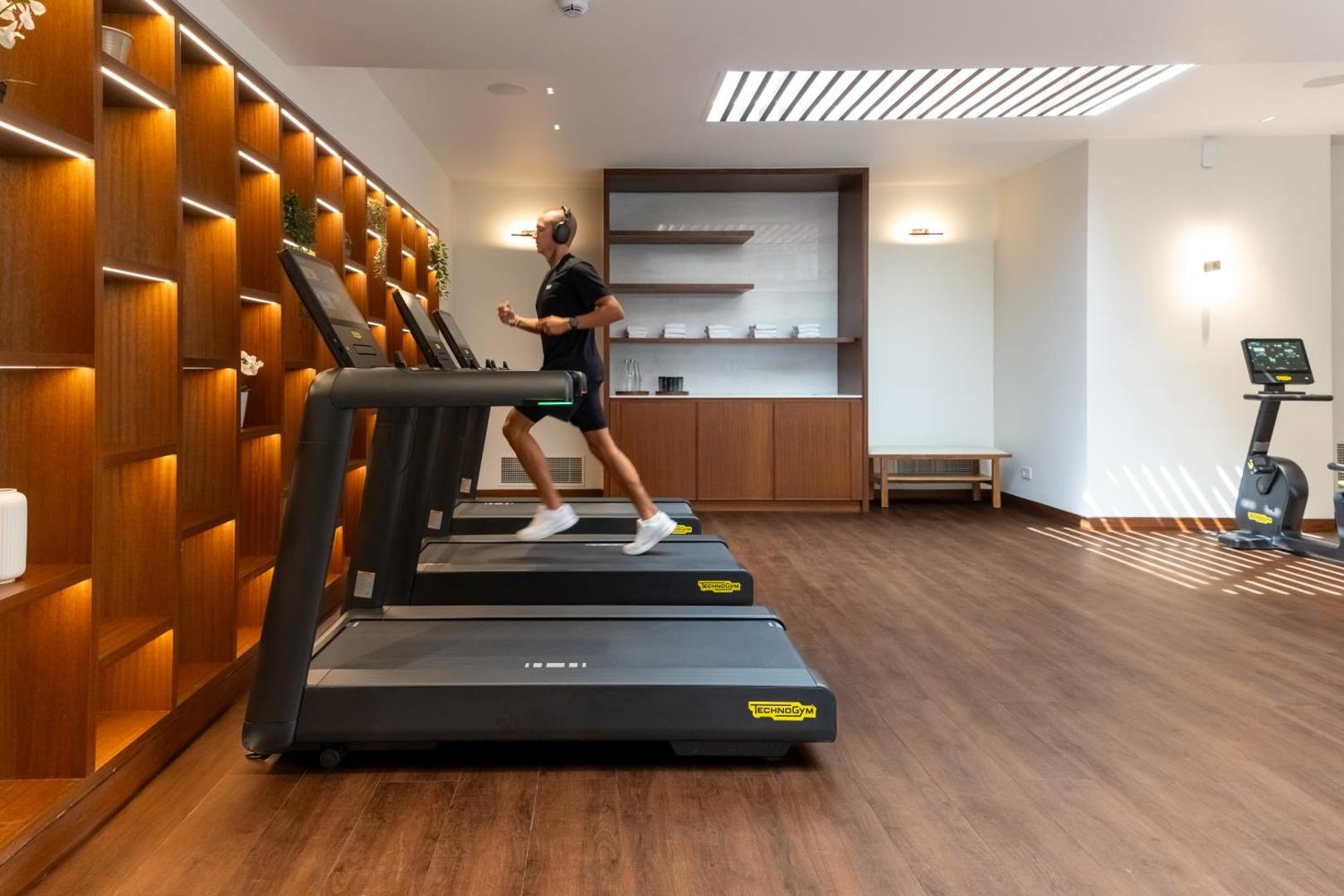 Viesis trenējas Porto Elounda Golf & Spa Resort sporta zālē uz skrejceļa — plaša telpa ar koka dizaina elementiem