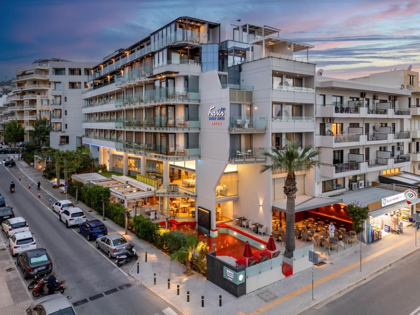 Kriti Beach Hotel fasāde vakarā Retimno pilsētā Krētā — moderna pieczvaigžņu viesnīca ar apgaismotiem balkoniem un āra terasi pie ielas