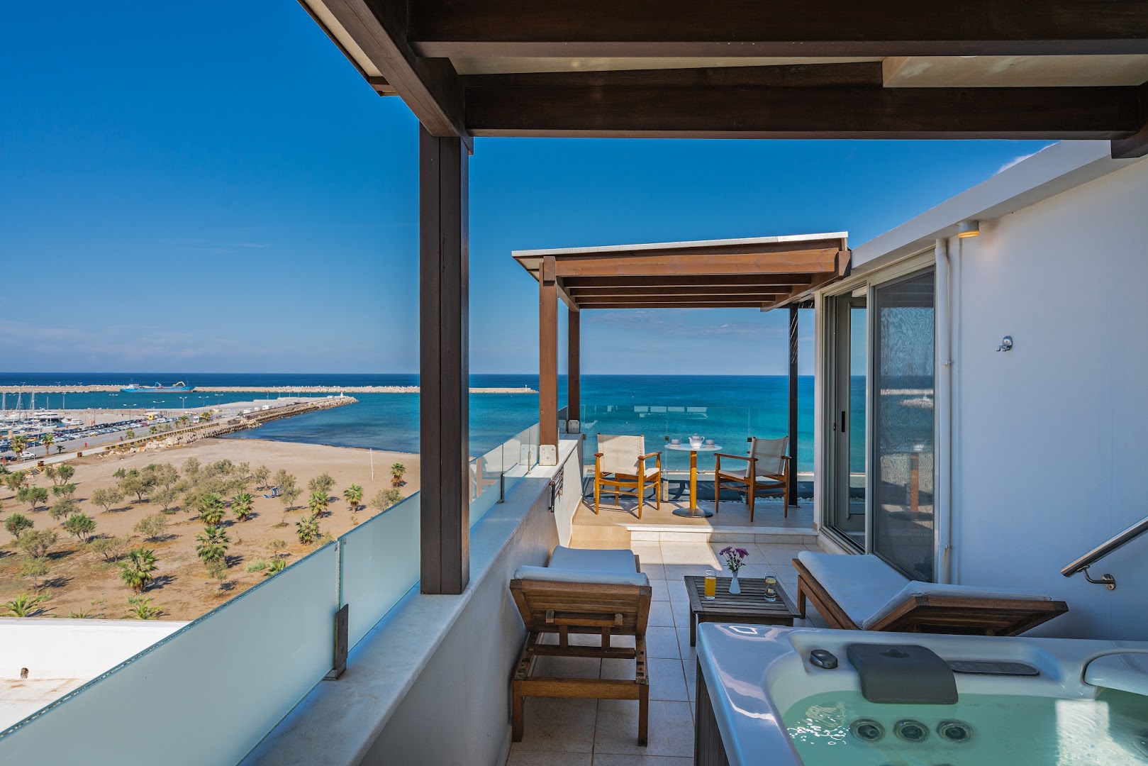 Kriti Beach Hotel luksusa numura terase ar džakuzi — koka nojume, sauļošanās krēsli un skats uz Retimno pludmali un ostu
