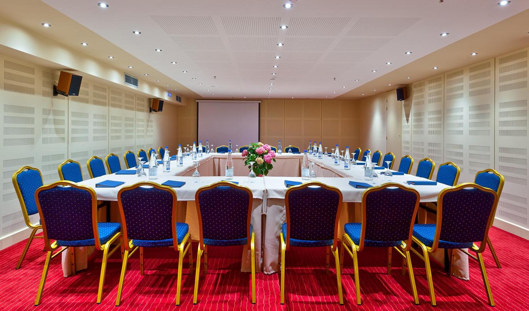 Kriti Beach Hotel konferenču zāle ar U-veida galda izkārtojumu, zilajiem krēsliem un prezentāciju aprīkojumu biznesa pasākumiem.