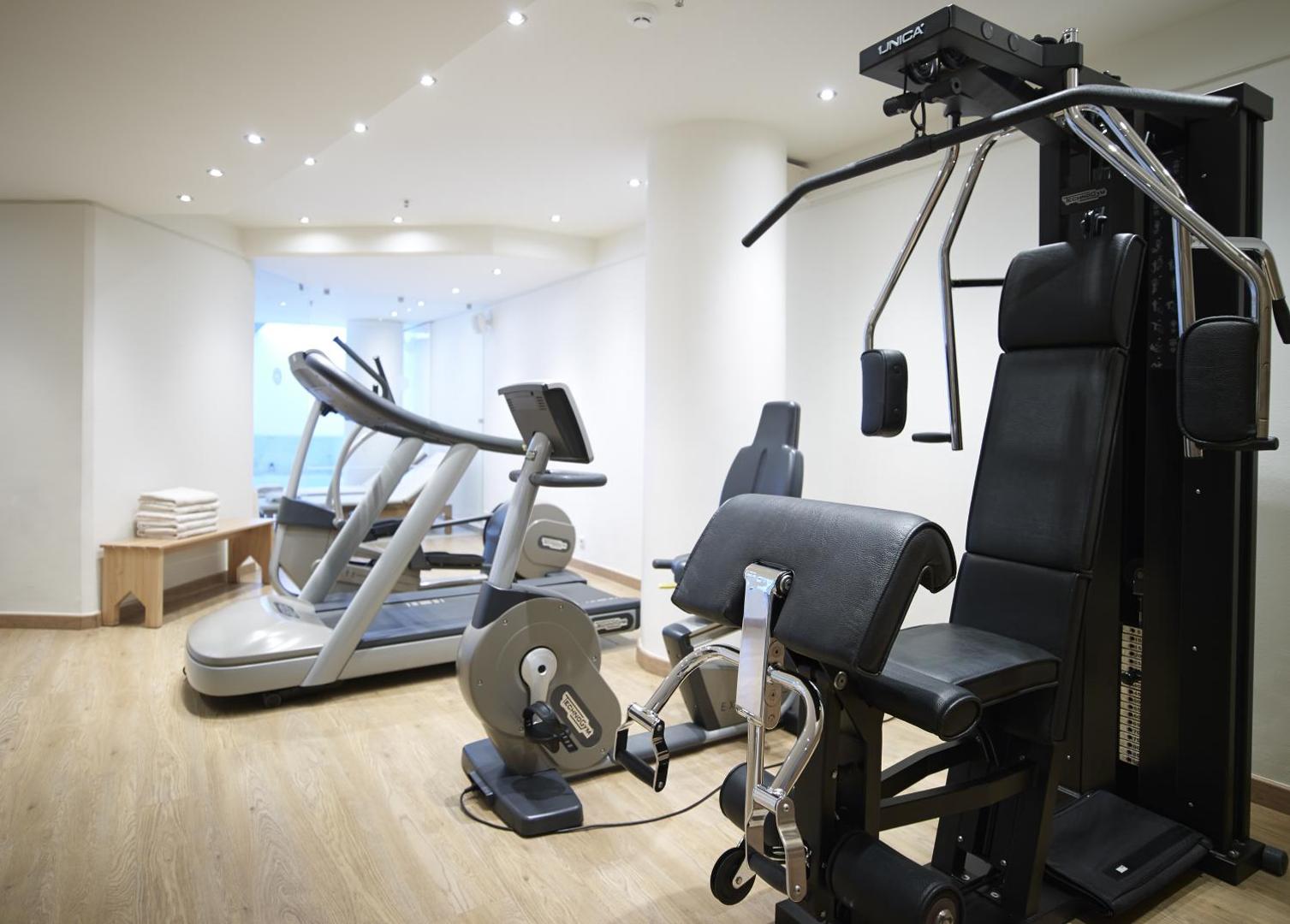 Kriti Beach Hotel fitnesa zāle ar Technogym kardio trenažieriem un spēka staciju — gaišs interjers ar koka grīdu un dvieļiem viesiem.