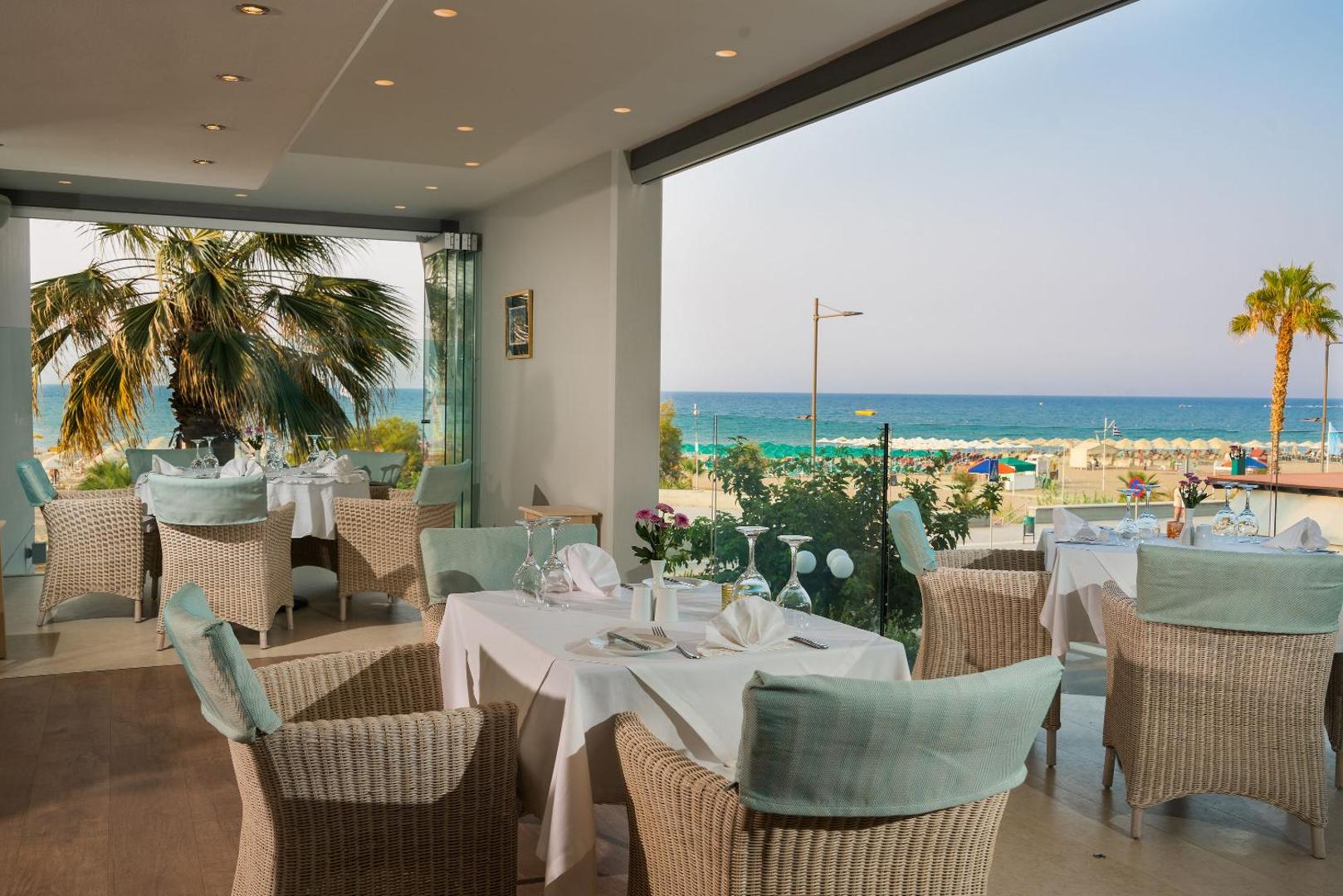 Kriti Beach Hotel gaišais restorāns ar panorāmas logiem uz pludmali un palmām — mājīgs interjers ar pītiem krēsliem un gaiši ziliem akcentiem