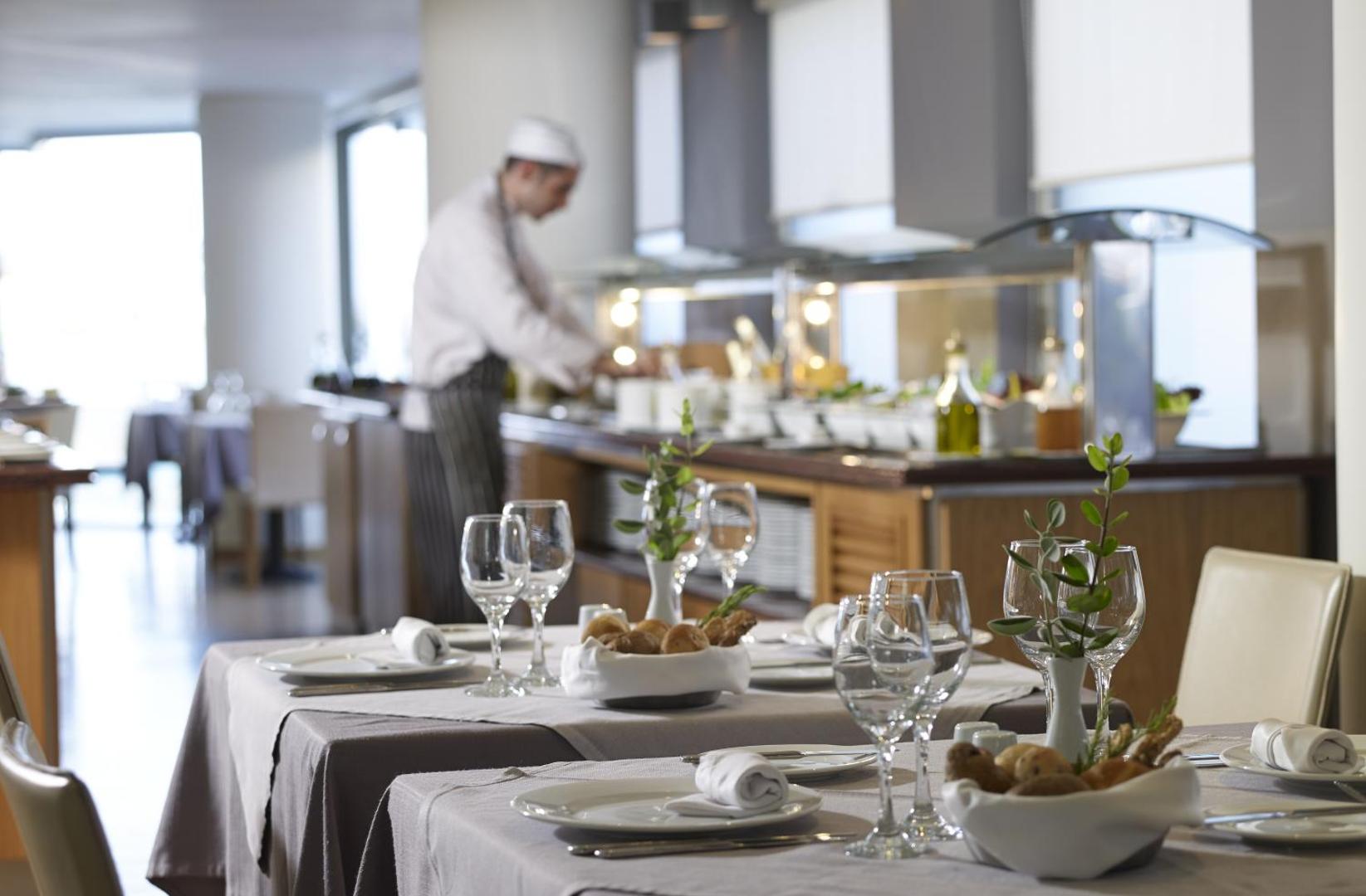 Kriti Beach Hotel bufetes tipa restorāns ar pavāru fonā un eleganti klātiem galdiem priekšplānā — svaiga maize un vīna glāzes