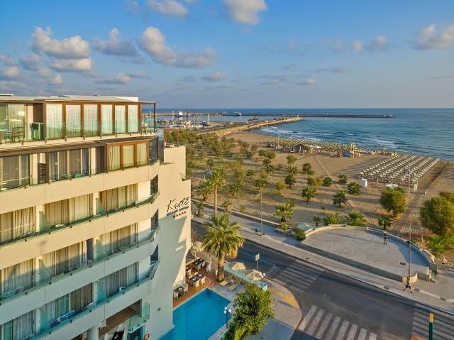Skats no augšas uz Kriti Beach Hotel Retimno — redzama viesnīcas ēka, āra baseins, iela un plašā smilšu pludmale pie jūras