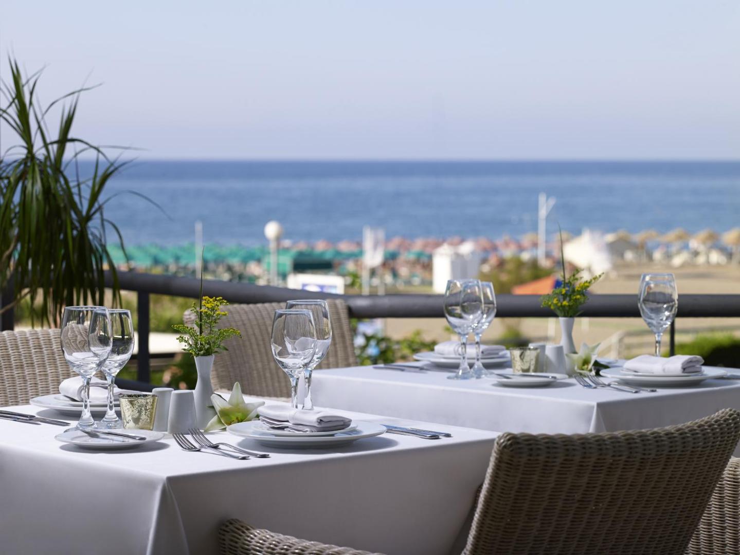 Kriti Beach Hotel restorāna terase ar klātiem galdiem un skatu uz Vidusjūru Retimno piekrastē, Krētā — eleganti balti galdauti un pīti krēsli