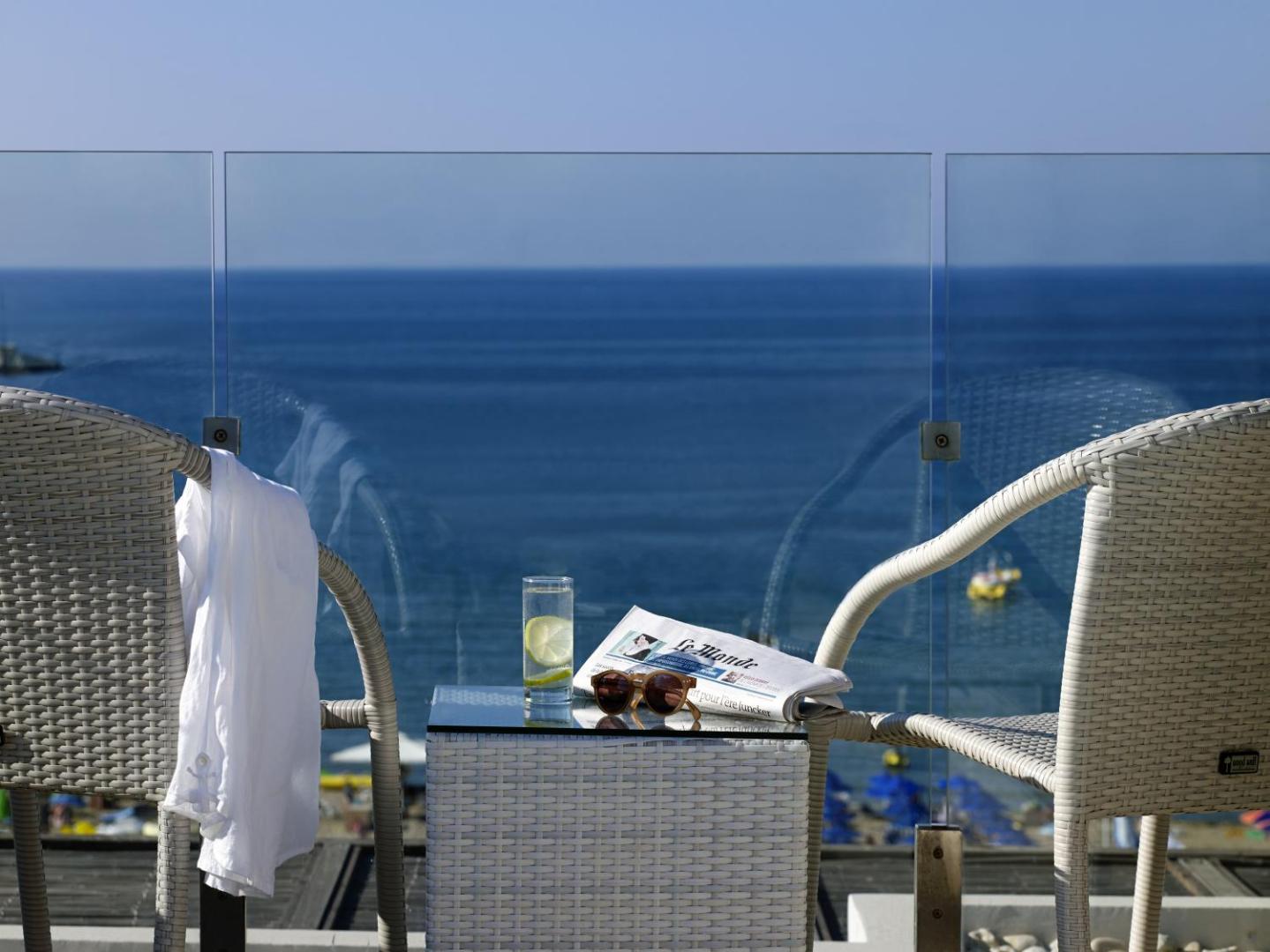 Kriti Beach Hotel balkons ar skatu uz Vidusjūru — pītie krēsli, galds ar dzērienu un avīzi pie stikla margām