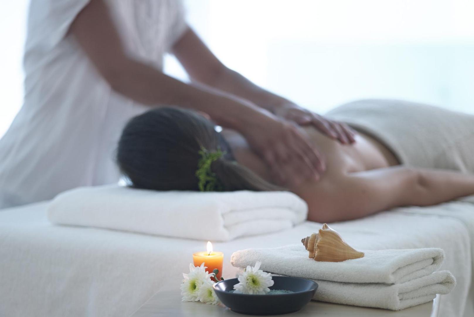 Relaksējoša masāža Kriti Beach Hotel spa centrā — sieviete guļ uz masāžas galda, priekšplānā svece, ziedi un dekoratīvs gliemežvāks