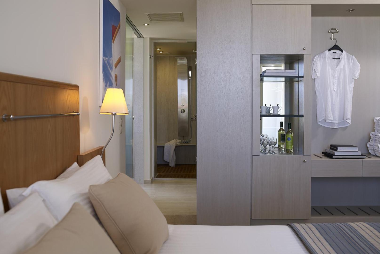 Kriti Beach Hotel numura interjers ar skatu uz vannas istabu — gaišas mēbeles, minibārs un mīksta gulta