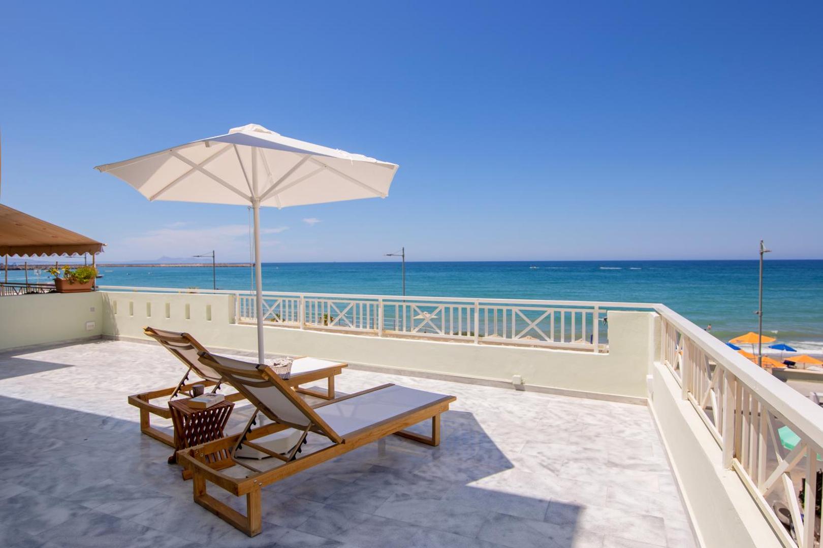 Ikones Seafront Luxury Suites plaša terase ar skatu uz jūru — koka sauļošanās krēsli, balts saulessargs un marmora grīdas segums