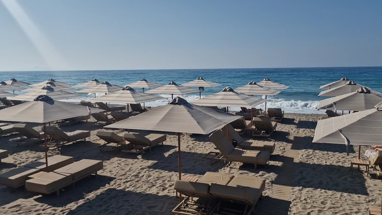 Ikones Seafront Luxury Suites pludmales zona ar daudziem saulessargiem un ērtiem zviļņiem gaišās smiltīs pie zilās jūras