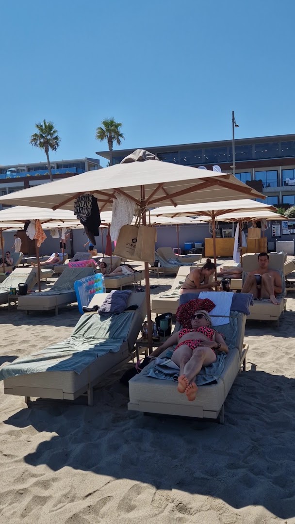 Ikones Seafront Luxury Suites viesi bauda sauli uz mīkstiem zviļņiem zem lieliem saulessargiem viesnīcas pludmalē