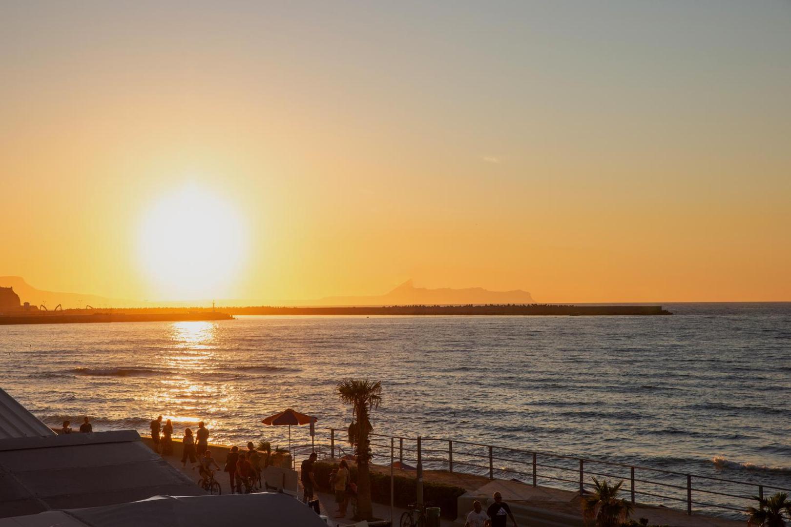 Ikones Seafront Luxury Suites skats uz saulrietu Retimno piekrastē Krētā — silta vakara gaisma pār jūru un promenādi ar palmām