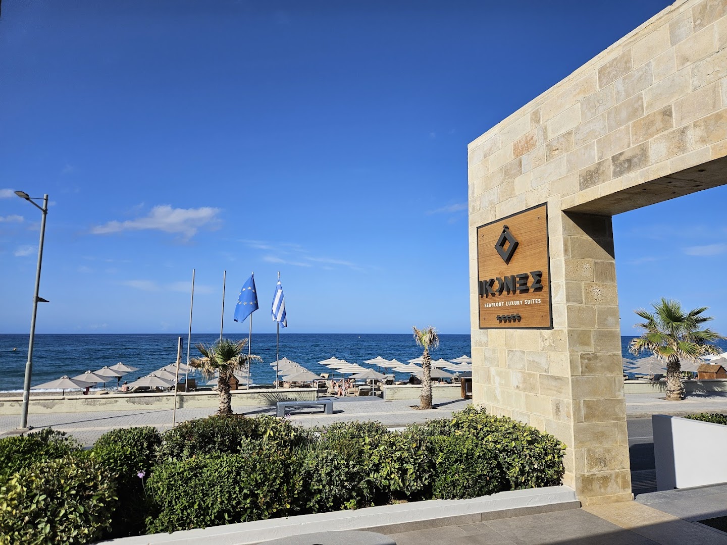 Ikones Seafront Luxury Suites ieeja pie pludmales Retimno — skats uz jūru, saulessargiem un zilajiem karogiem pie viesnīcas zīmes
