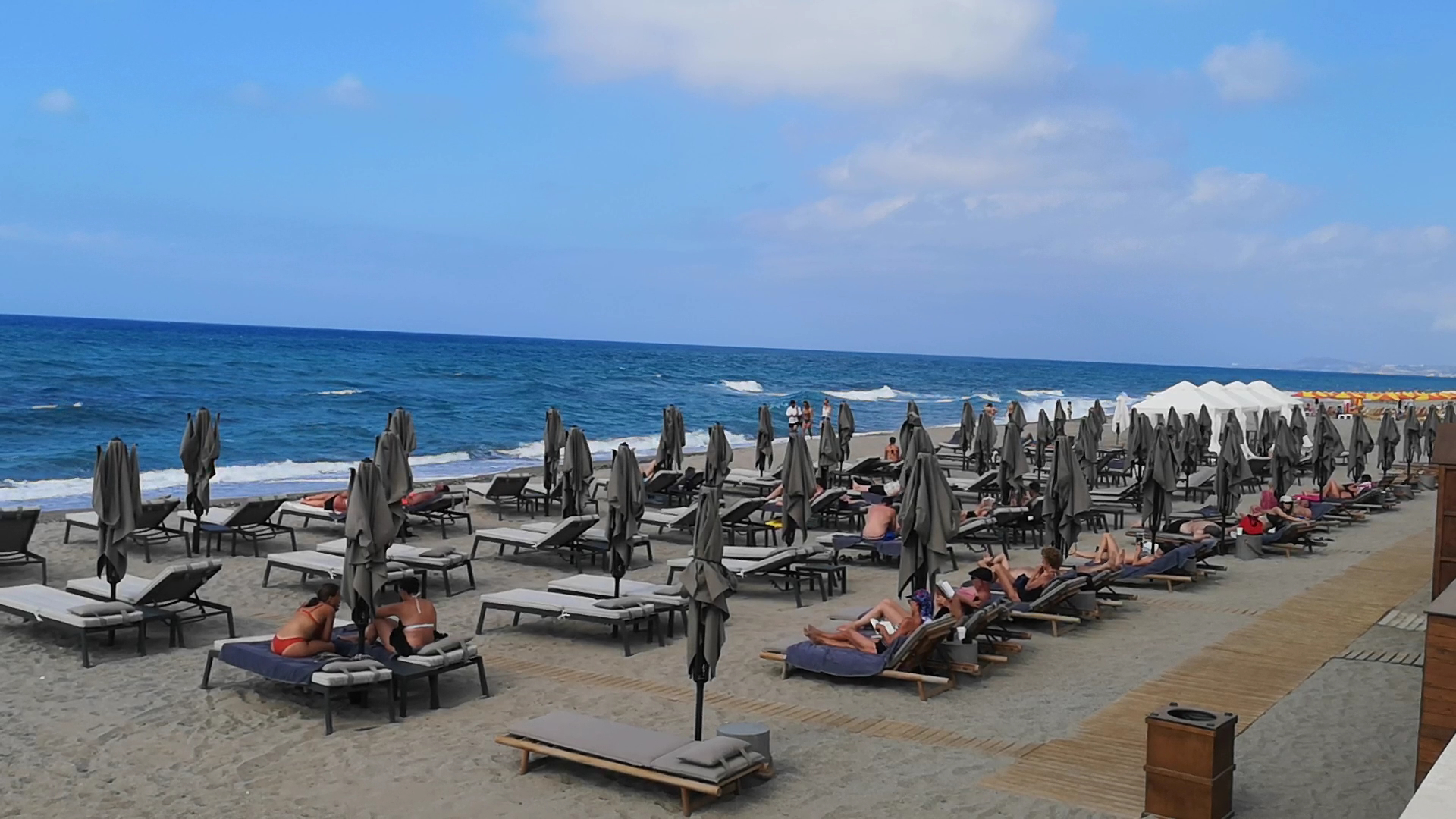 Ikones Seafront Luxury Suites pludmales skats ar viesiem, kuri atpūšas uz zviļņiem pie jūras krasta līnijas