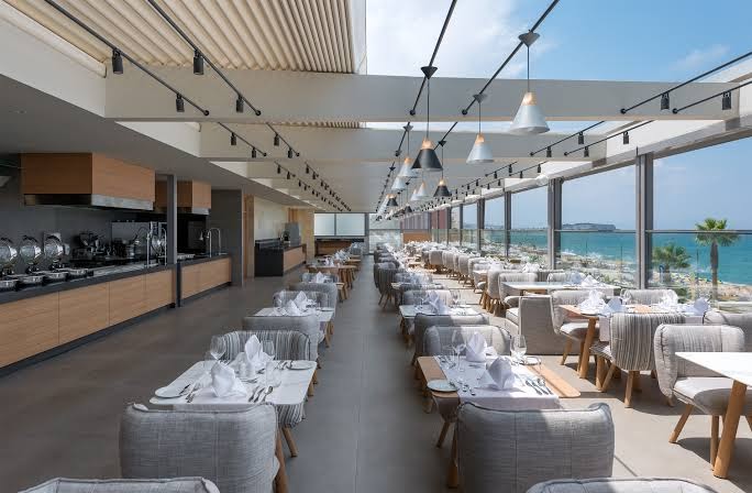 Ikones Seafront Luxury Suites restorāns ar skatu uz jūru Retimno, Krētā — eleganti klāti galdi, moderna bufetes zona un panorāmas logi
