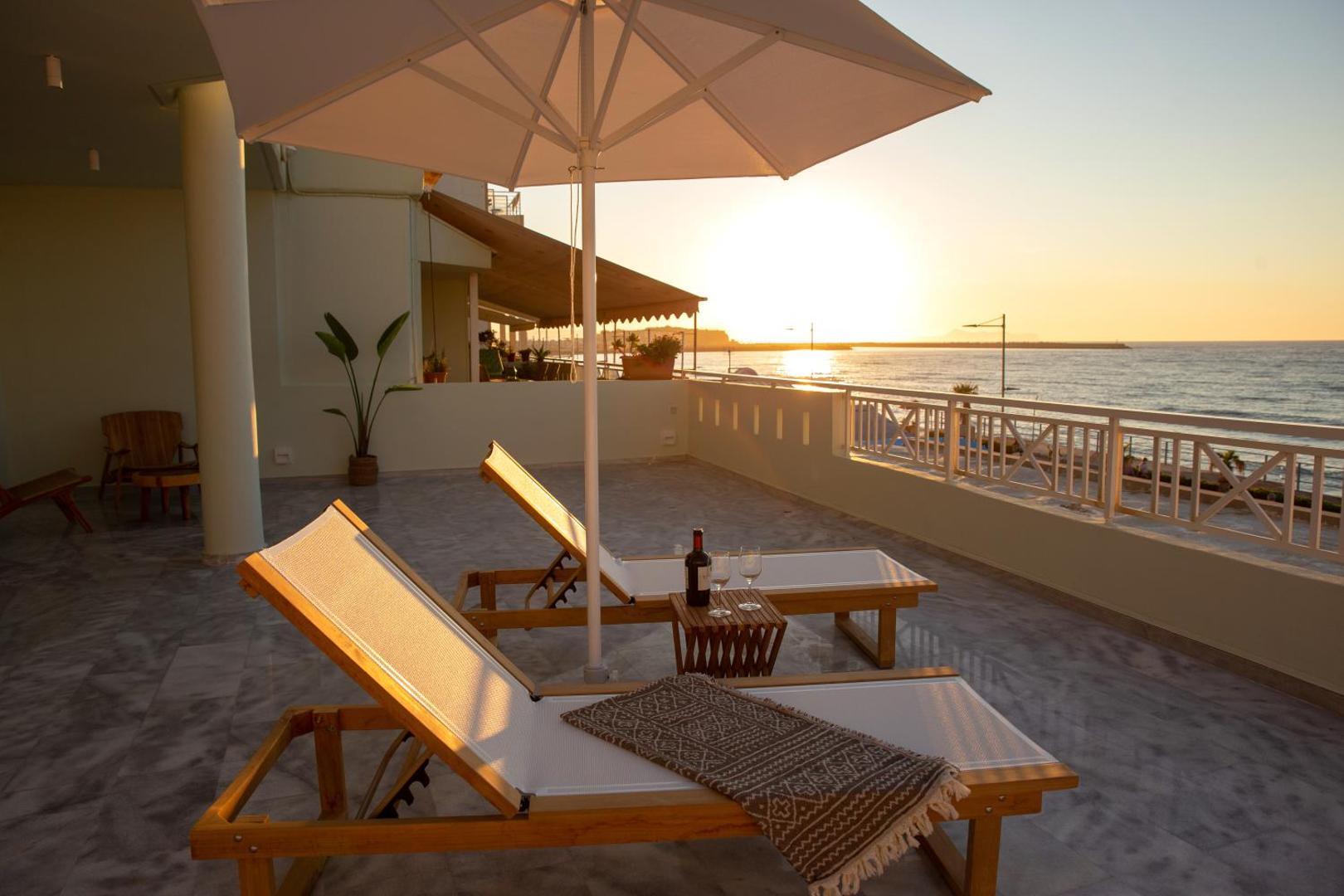 Ikones Seafront Luxury Suites terase ar sauļošanās krēsliem un skatu uz jūru saulrietā Retimno, Krētā — romantiska gaisotne ar vīnu