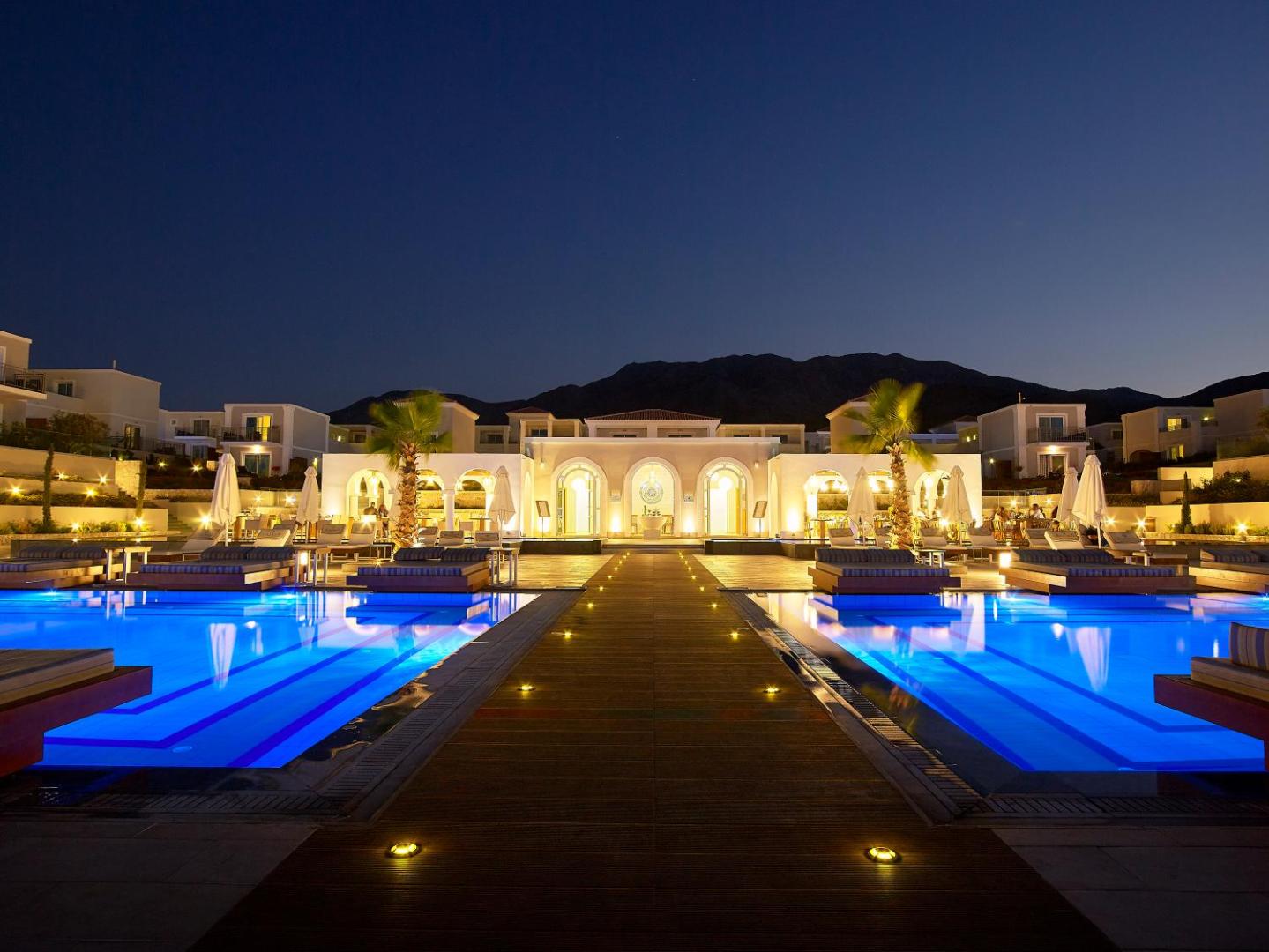 Anemos Luxury Grand Resort centrālā ieeja un baseini nakts apgaismojumā ar kalnu siluetu fonā Krētas piekrastē