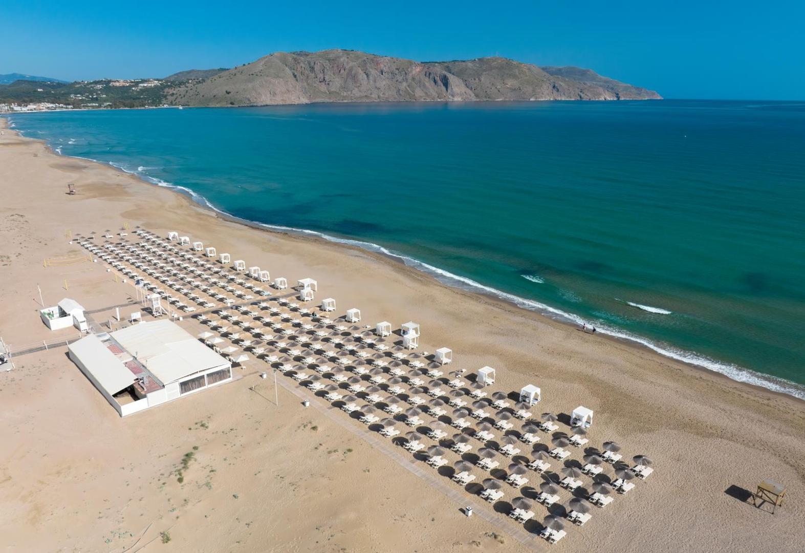 Anemos Luxury Grand Resort plašā smilšu pludmale ar saulessargiem un gultām pie dzidri zilās jūras Krētā