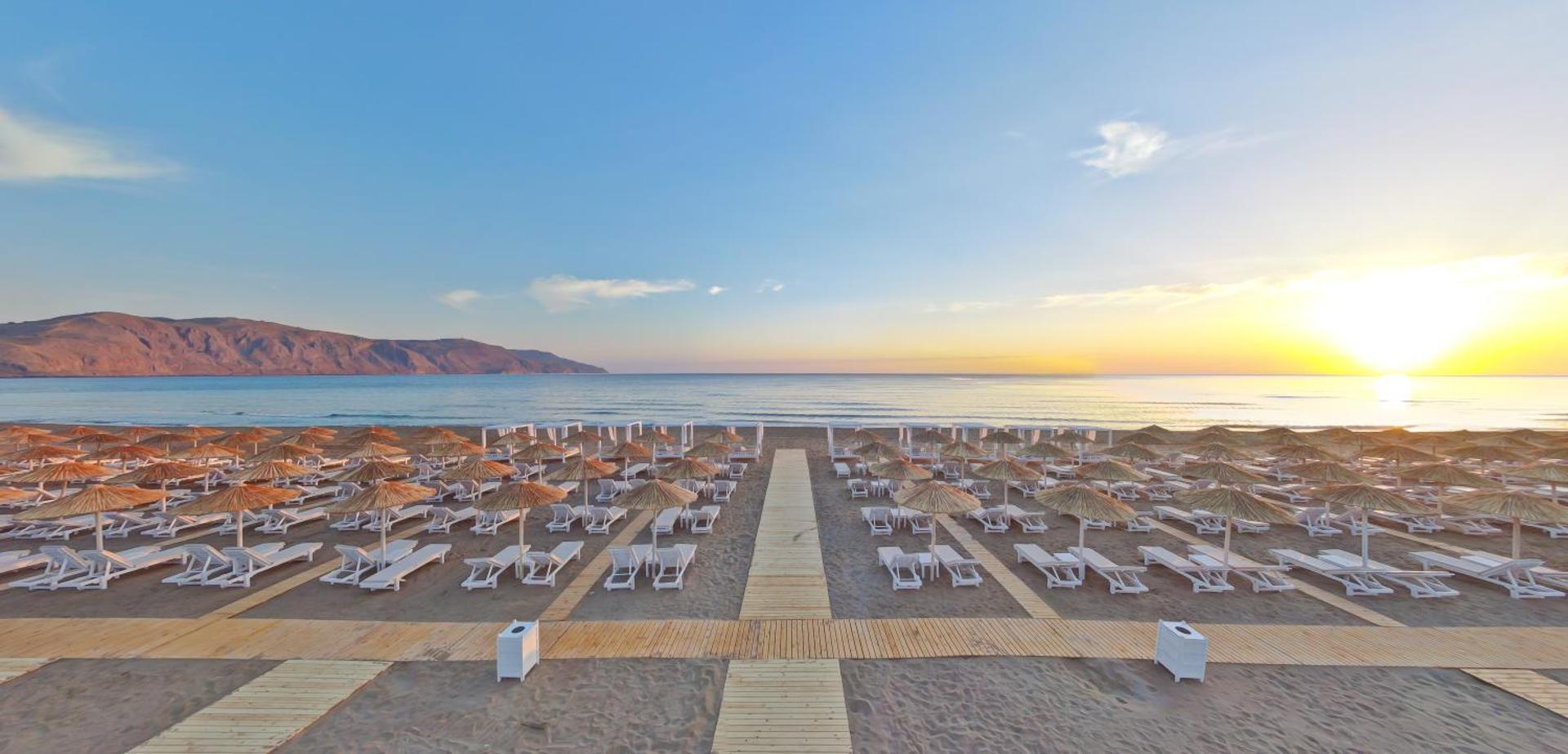 Anemos Luxury Grand Resort plašā smilšu pludmale saulrietā Georgiupoli, Krētā — sakārtotas saulessargu rindas un koka laipas pie jūras