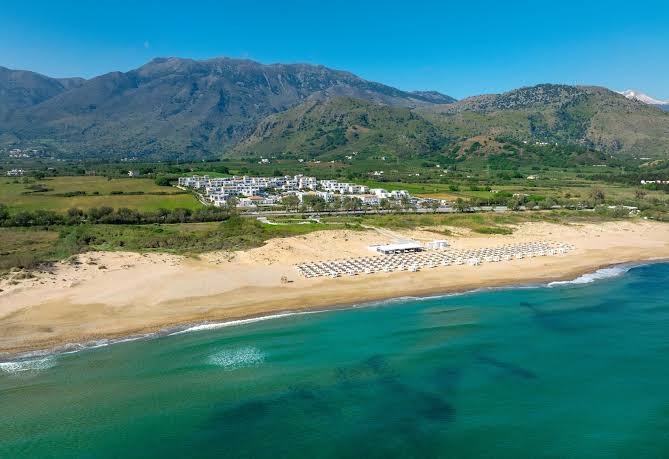 Aerofoto skats uz Anemos Luxury Grand Resort plašo smilšu pludmali, zilo jūru un kalniem fonā Krētā