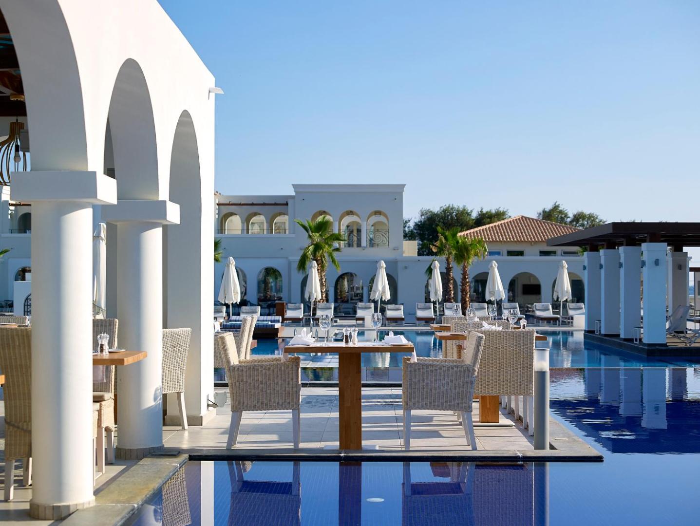 Anemos Luxury Grand Resort āra terase pie baseina ar elegantiem galdiņiem un baltām arkām uz viesnīcas arhitektūras fona