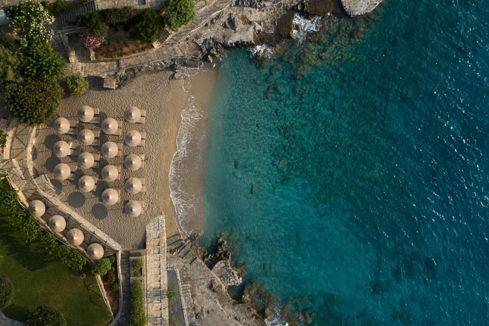Minos Palace Resort privātā smilšu pludmale no putna lidojuma — saulessargi, atpūtas krēsli un kristāldzidrs tirkīza krāsas ūdens