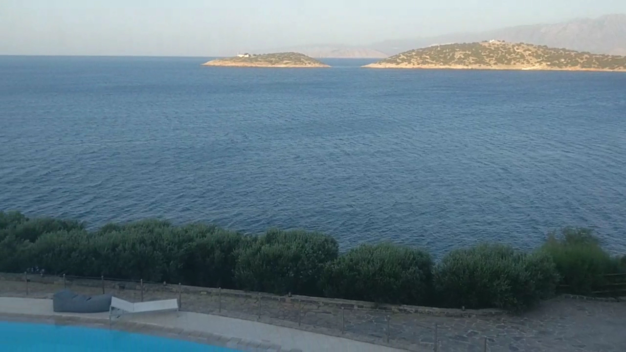 Skats no Minos Palace Resort numura terases uz jūru un divām salām līcī — baseina mala un atpūtas krēsli priekšplānā