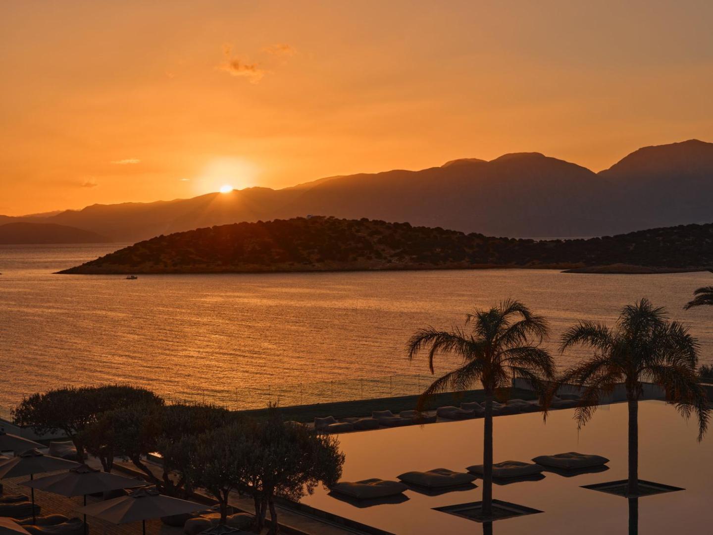 Minos Palace Resort skats uz saulrietu virs Mirabello līča — panorāmas baseins ar palmām un kalnu siluetu fonā Krētā