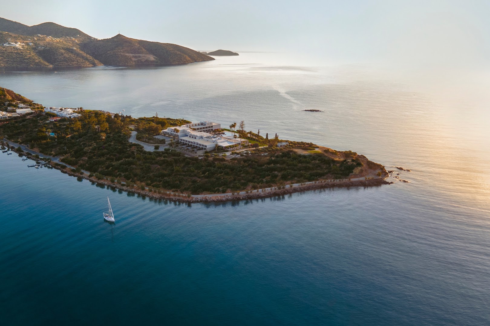 Minos Palace Resort skats no putna lidojuma saulrietā — viesnīcas ēkas uz pussalas kailu kalnu un rāmas jūras ielokā