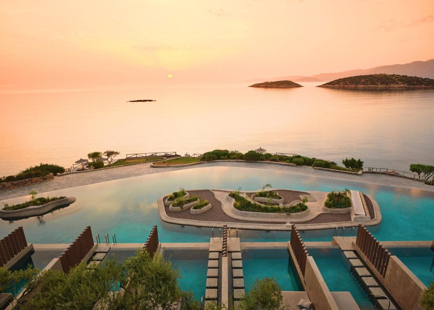 Minos Palace Resort panorāmas skats uz baseinu kompleksu, privātajiem baseiniem un jūras līci saulrietā