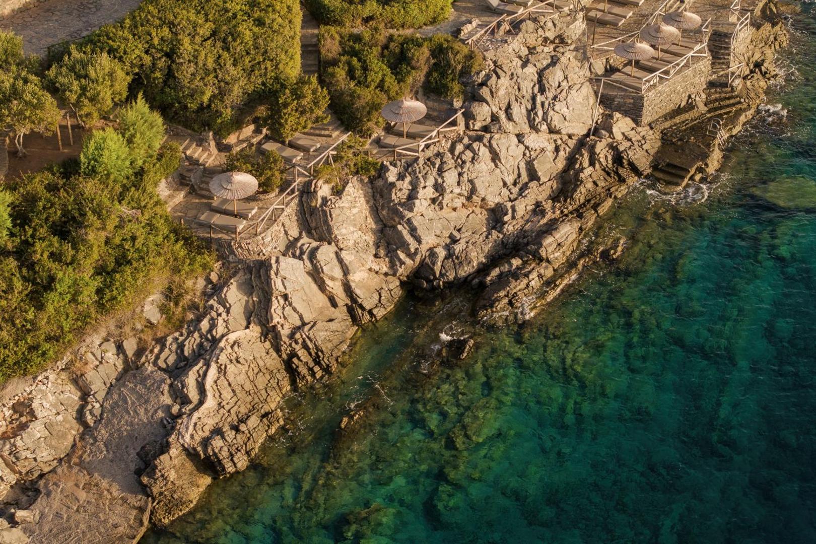 Minos Palace Resort klinšainā piekraste ar privātām sauļošanās platformām, saulessargiem un kāpnēm uz dzidro jūras ūdeni