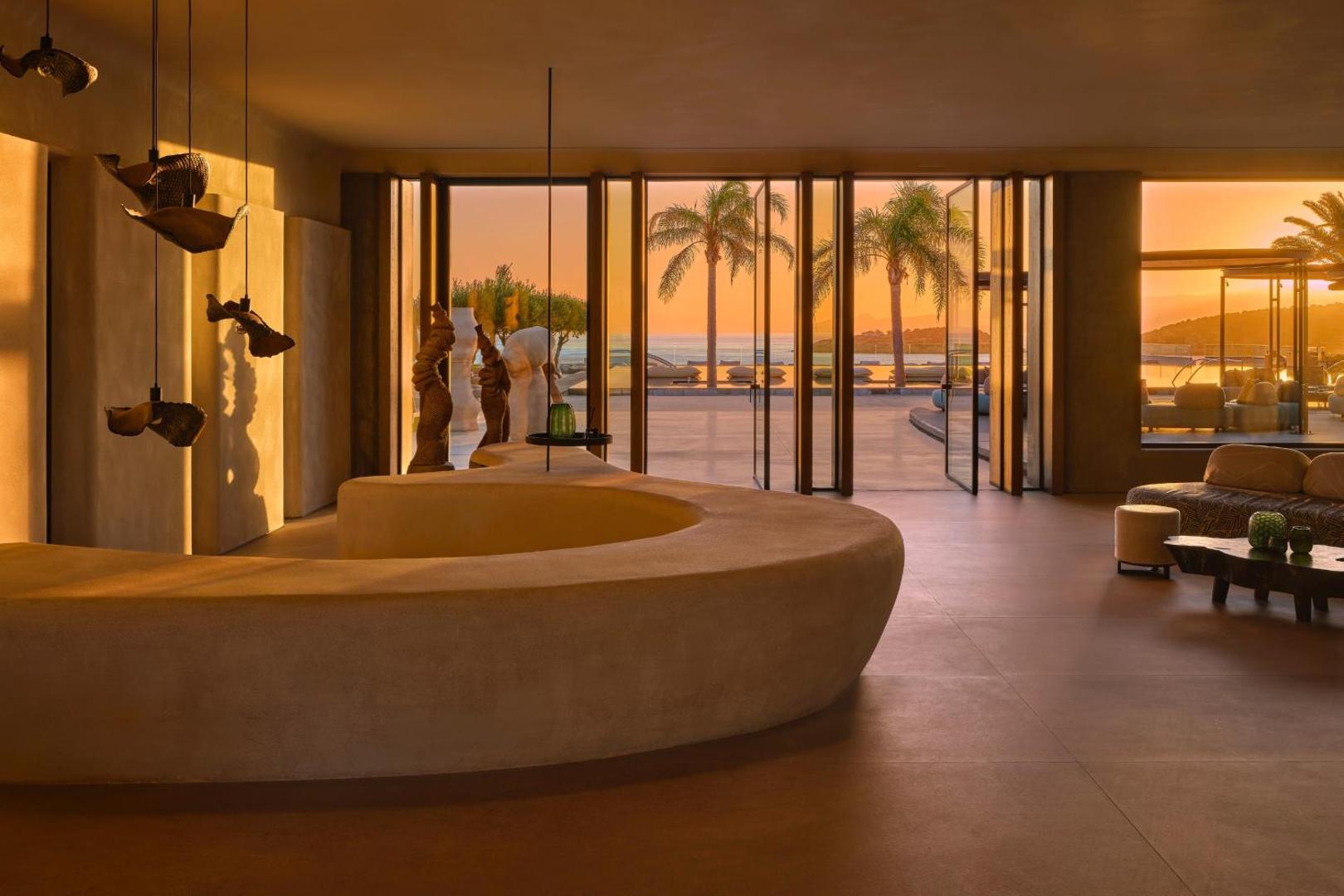 Minos Palace Resort lobby zona saulrieta laikā ar minimālistisku dizainu, skulpturālām formām un skatu uz palmām.