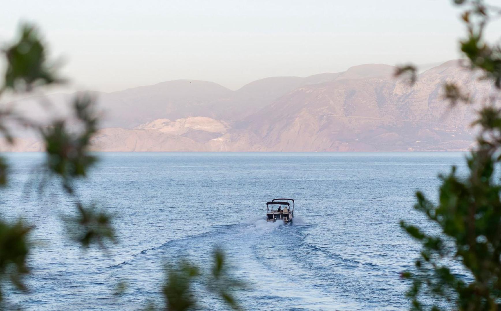 Motorlaiva jūrā pie Elounda Mare Hotel, skats caur koku zariem uz kalnaino Krētas piekrasti saulrietā