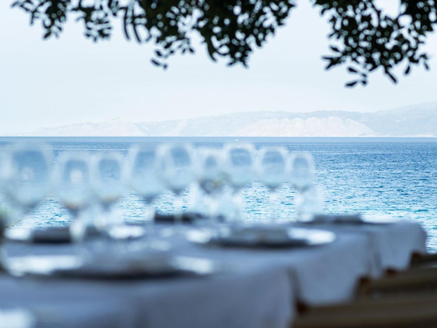 Skats uz Egejas jūru un kalniem no Elounda Mare Hotel restorāna galda — fokusā dzidras vīna glāzes uz balta galdauta