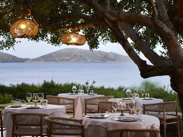 Elounda Mare Hotel restorāns zem koku zariem ar skatu uz Mirabello līci — eleganti klāti galdi un pīti gaismekļi vakarā