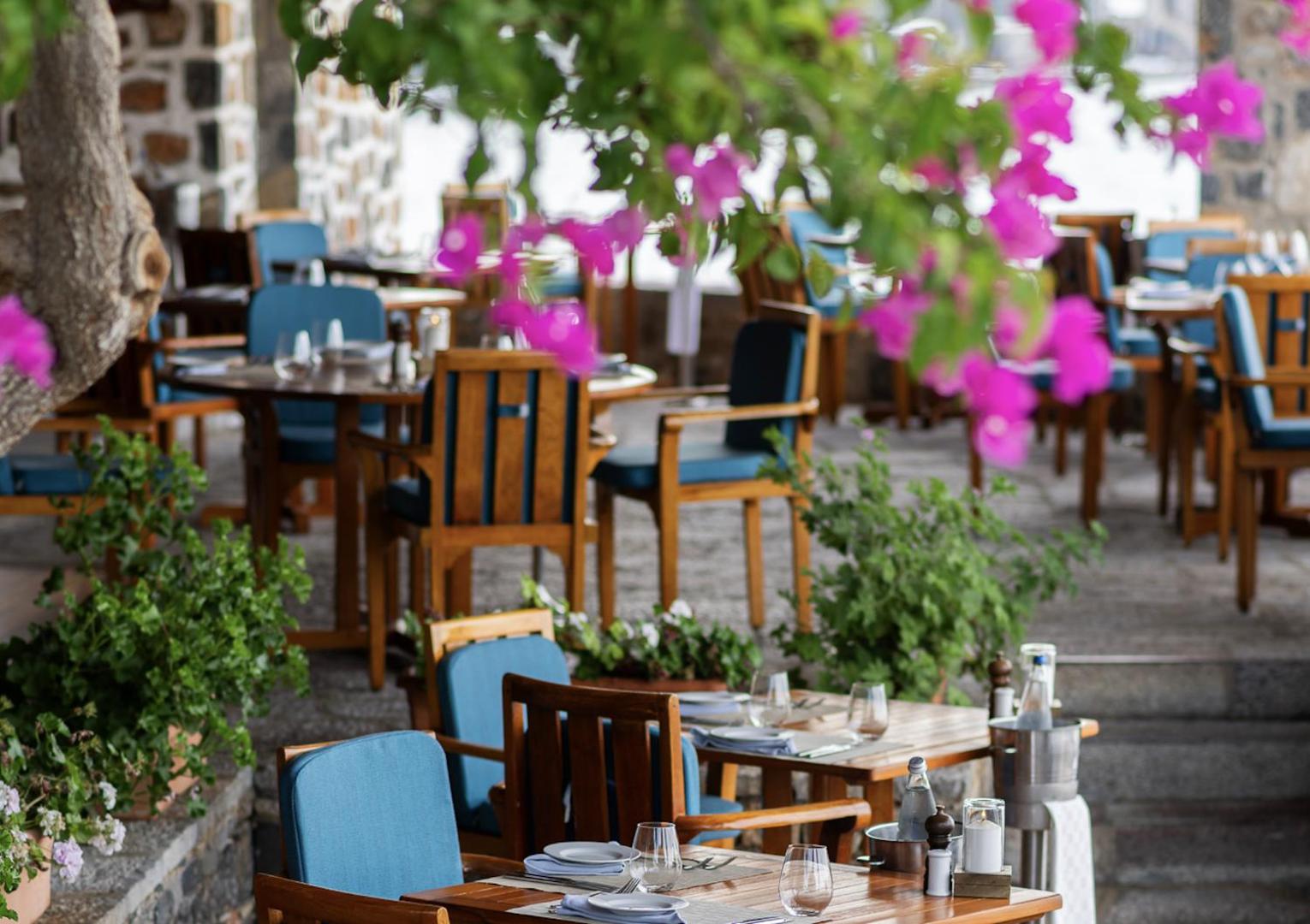 Elounda Mare Hotel āra restorāna terase ar koka mēbelēm un ziliem akcentiem, ko ieskauj ziedošas bugenvilijas un akmens sienas