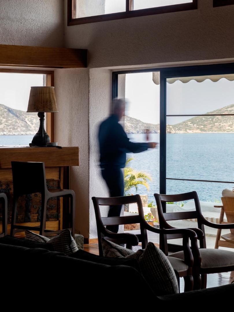 Skats no Elounda Mare Hotel atpūtas telpas uz jūru caur lielu logu, redzami koka krēsli un piekrastes ainava