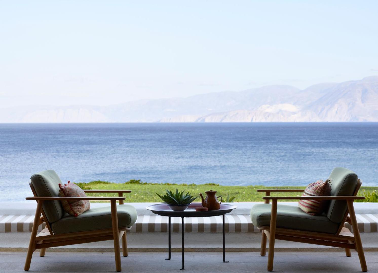 Terase ar ērtiem krēsliem un galdiņu Elounda Mare Hotel viesnīcā, fonā plašs skats uz jūru un kalnu grēdām