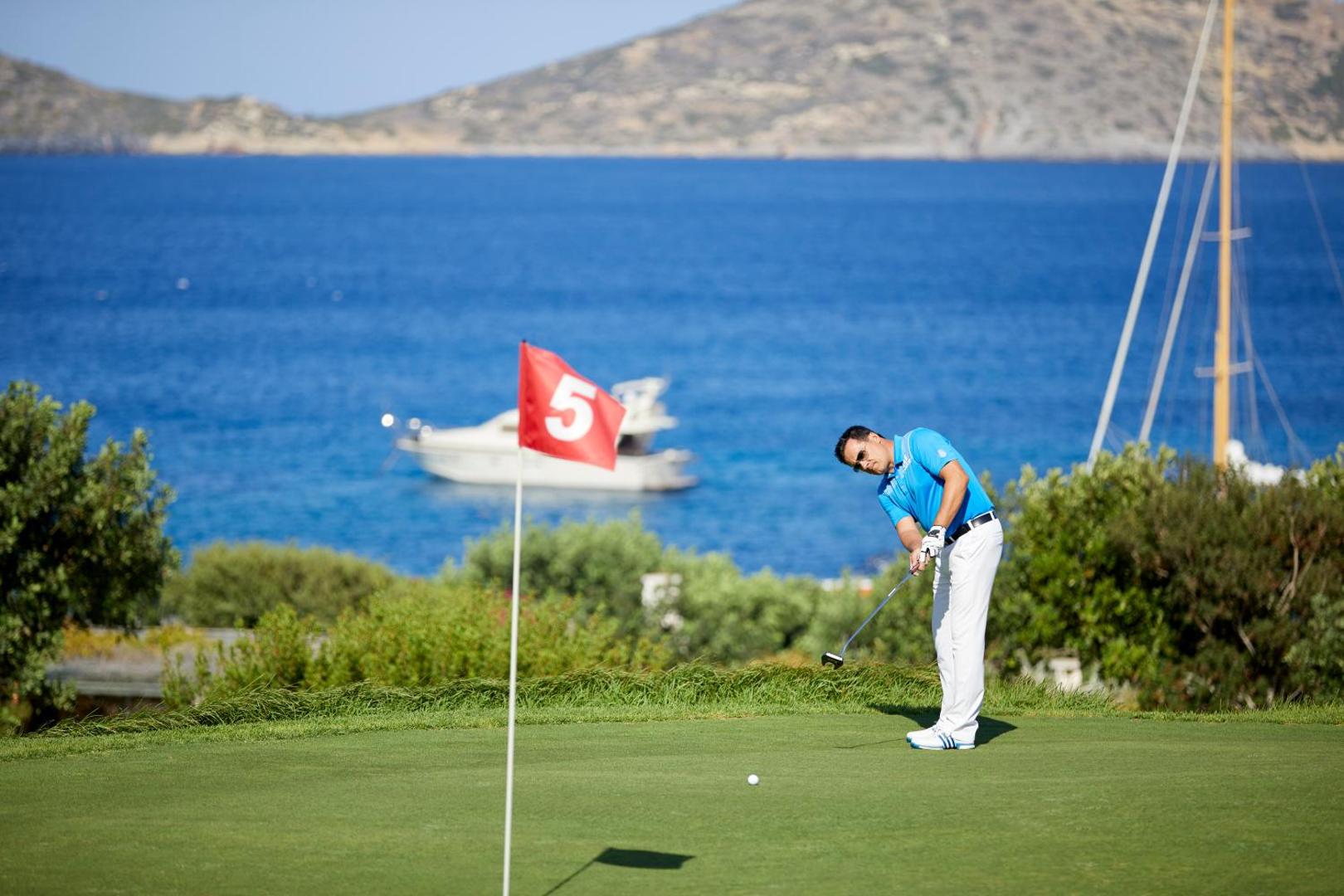 Golfa spēlētājs Elounda Mare Hotel laukumā pie jūras, fonā redzami kalni un jahtas zilā ūdenī
