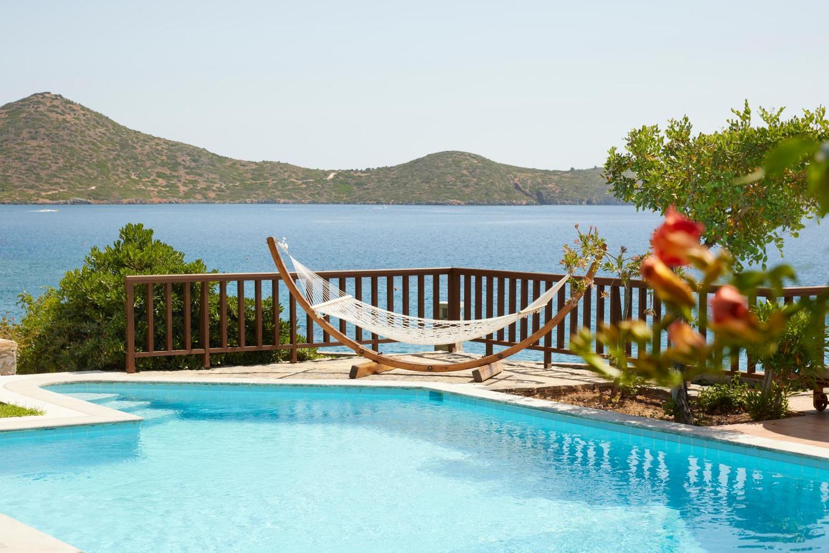 Elounda Mare Hotel baseina zona ar šūpuļtīklu un ziediem priekšplānā, fonā redzami Krētas kalni un jūra