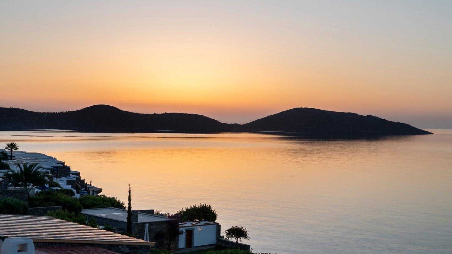 Elounda Mare Hotel skats uz Mirabello līci saulrieta laikā Krētā — mierīgs jūras ūdens un kalnu silueti pie horizonta