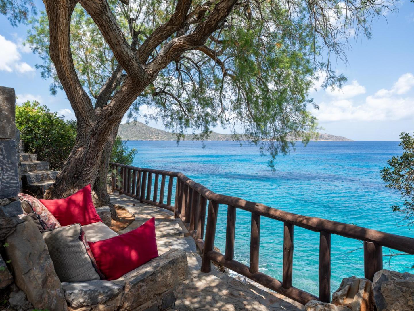Elounda Mare Hotel atpūtas zona piekrastē zem koka zariem ar skatu uz tirkīzzilo Vidusjūru un kalniem