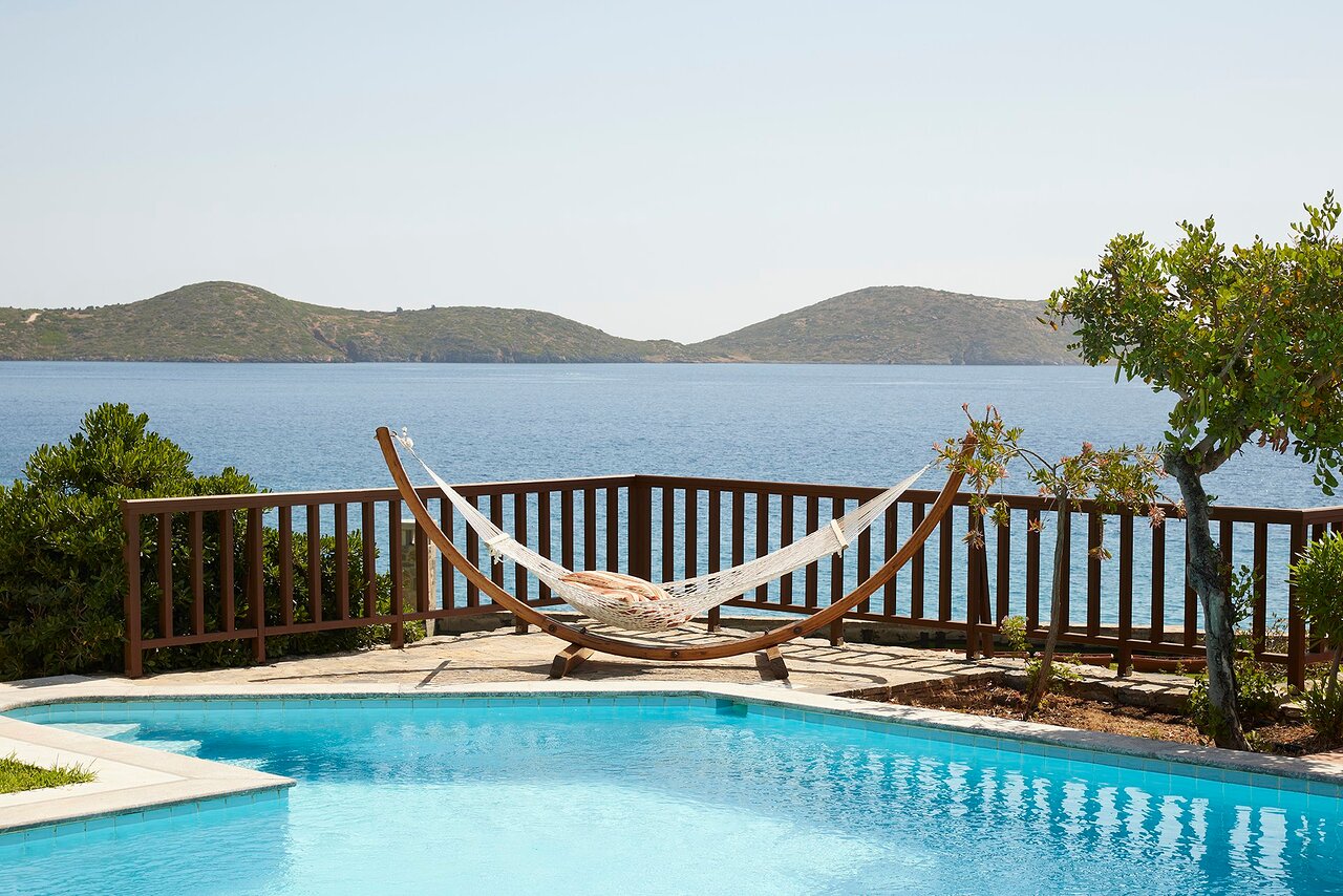 Elounda Mare Hotel atpūtas zona ar šūpuļtīklu pie baseina un skatu uz Mirabello līča zilo ūdeni un kalniem