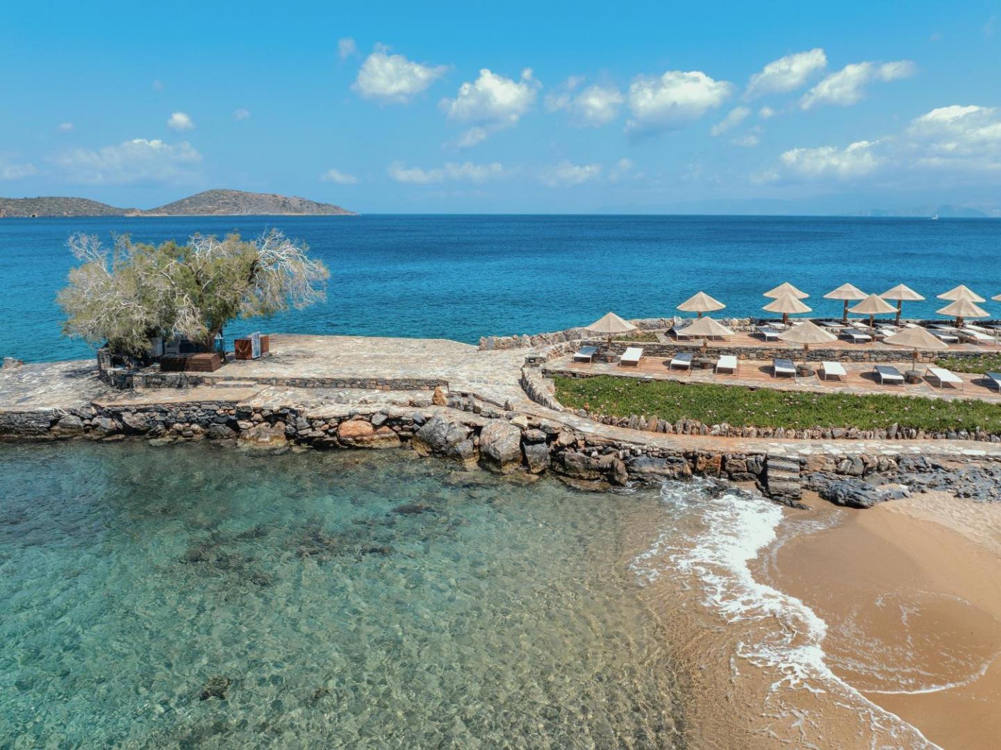 Elounda Mare Hotel atpūtas zona pie jūras — sauļošanās krēsli uz akmens terases un ieeja smilšu pludmalē
