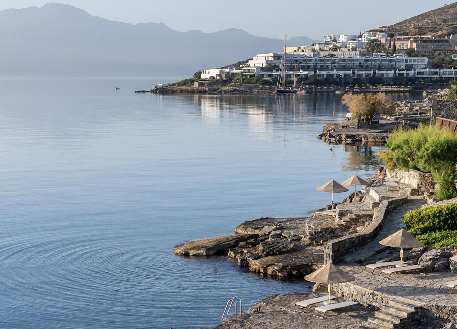 Elounda Mare Hotel akmeņainā pludmales zona ar saulessargiem un zviļņiem mierīgā rītā pie Mirabello līča