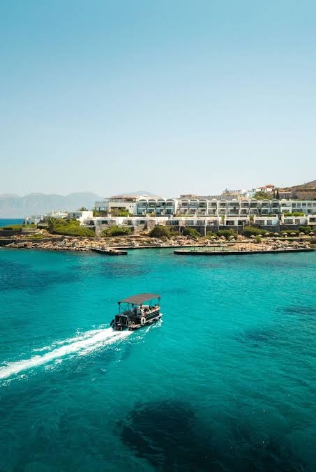 Skats no jūras uz Elounda Mare Hotel kompleksu un motorlaivu, kas pārvietojas pa tirkīzzilo ūdeni Krētā