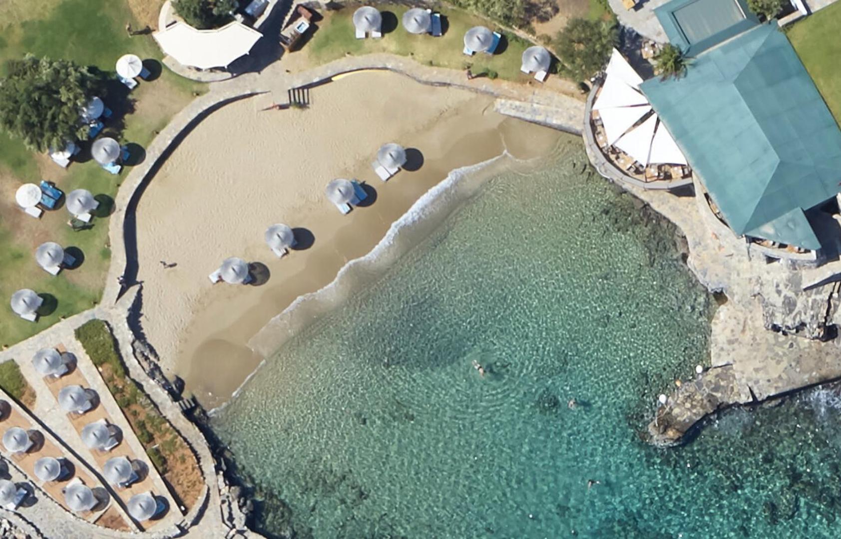 Aerofoto skats uz Elounda Mare Hotel smilšu pludmali — saulessargi, dzidrs ūdens un piekrastes restorāns no putna lidojuma
