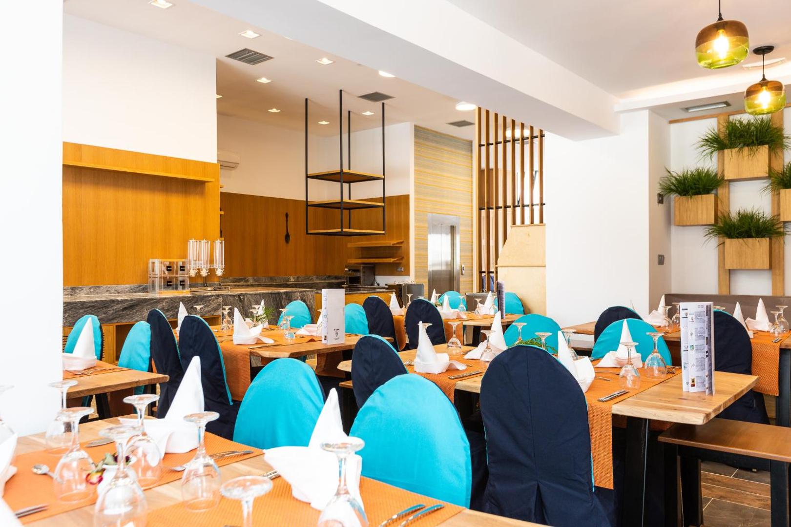 T Hotel Premium Suites plašā restorāna zāle ar servētiem galdiem, ziliem krēslu pārvalkiem un bufetes zonu fonā
